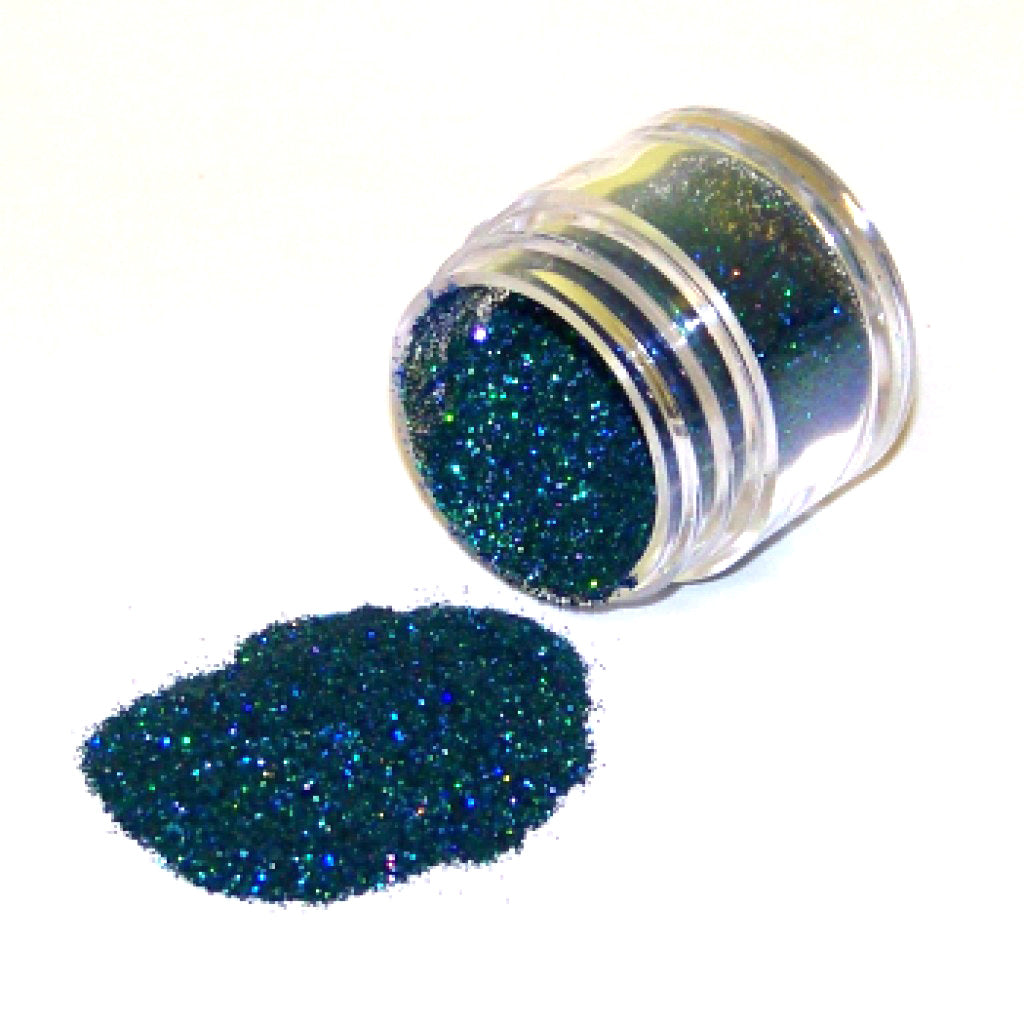 Small clear jar tipped on its side spilling peacock-colored glitter dust onto a white surface, with fine teal and blue sparkling particles visible throughout.