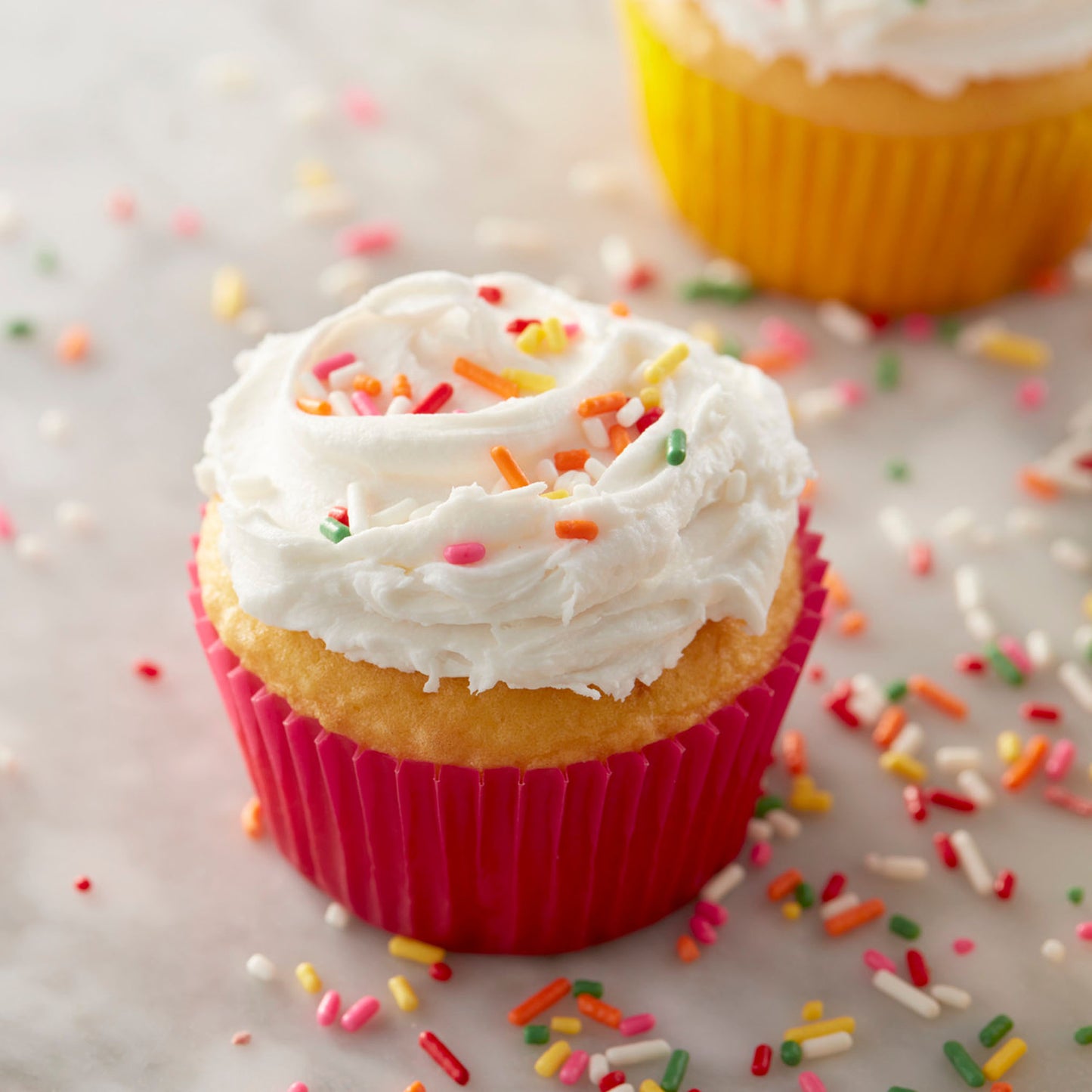 Frosted vanilla cupcake with sprinkles in a red cupcake liner, with yellow liner in background