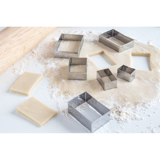 Six rectangular stainless steel cookie cutters on rolled dough with cut-out shapes and a rolling pin in view.