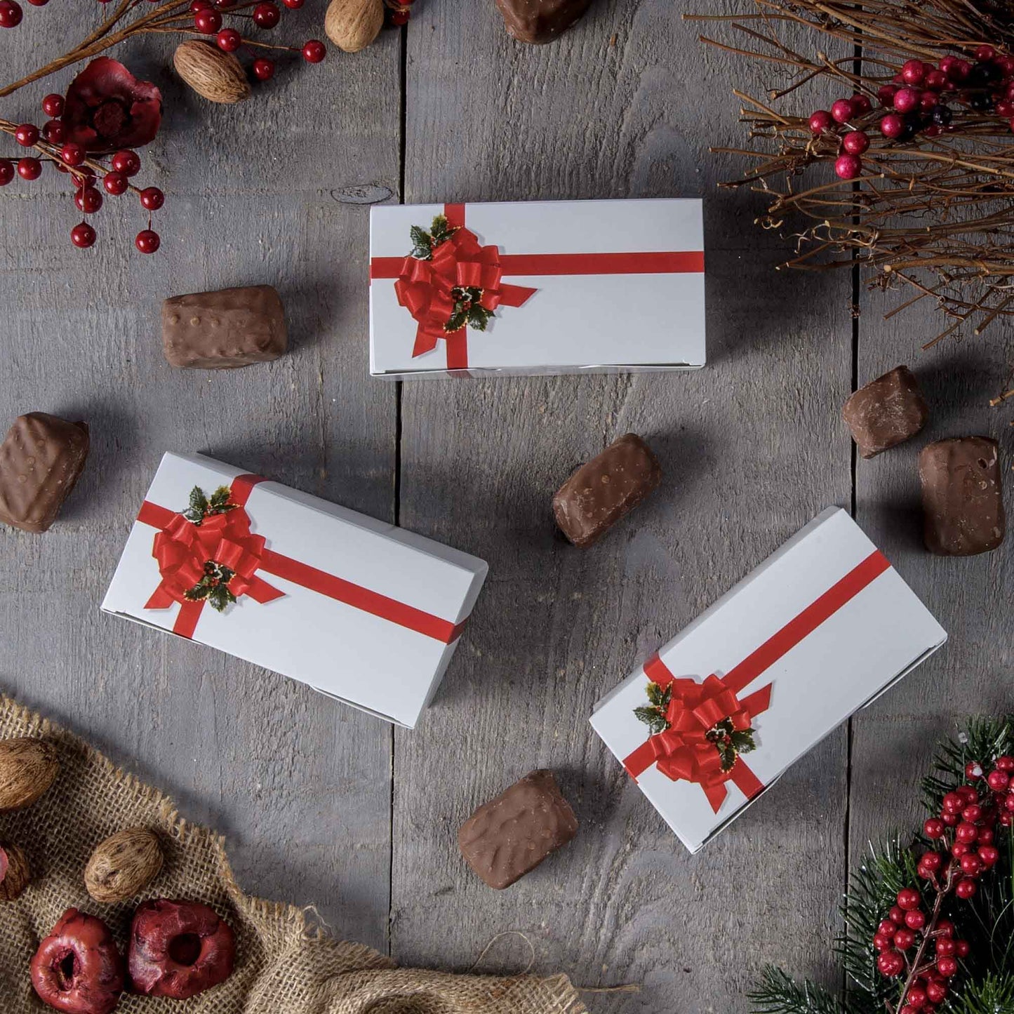 Red ribbon and holly candy boxes styled on a rustic wood surface with scattered chocolates, berries, and holiday greenery.
