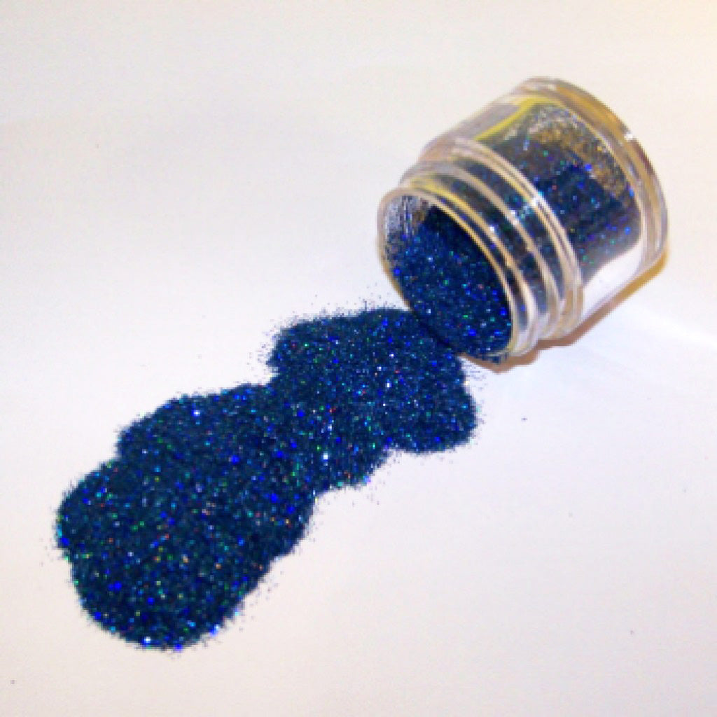 Small clear jar tipped on its side spilling sapphire blue glitter dust onto a white surface, with fine sparkling particles visible throughout.
