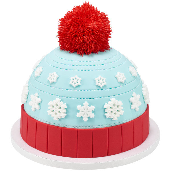 Winter hat-themed cake decorated with multiple white pressed sugar snowflakes on blue fondant and a large red pom-pom on top.