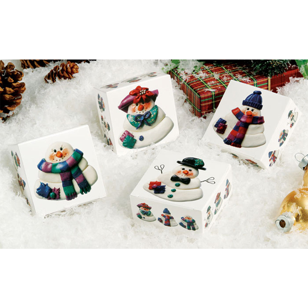 Four small square snowman-themed candy boxes displayed on white faux snow, each box showing a different snowman illustration with scarves and hats, with holiday decor in the background.