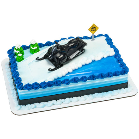 Rectangular winter-themed cake with blue piping and a snowy white top, decorated with a black snowmobile topper and a yellow snowmobile crossing sign.
