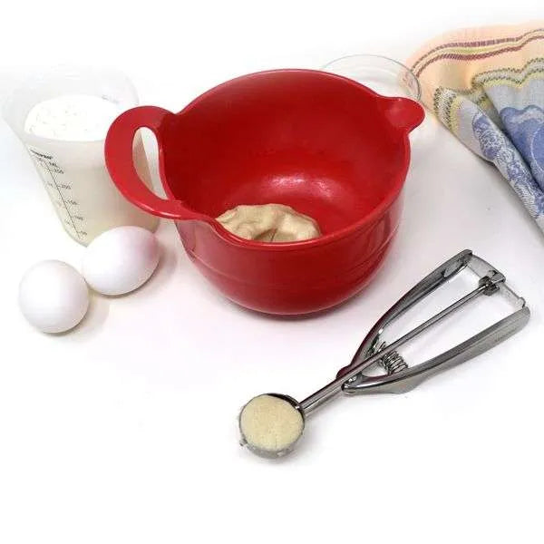 Baking scene with a red mixing bowl, flour, eggs, and a cookie scoop filled with sugar, showcasing ingredient portioning.