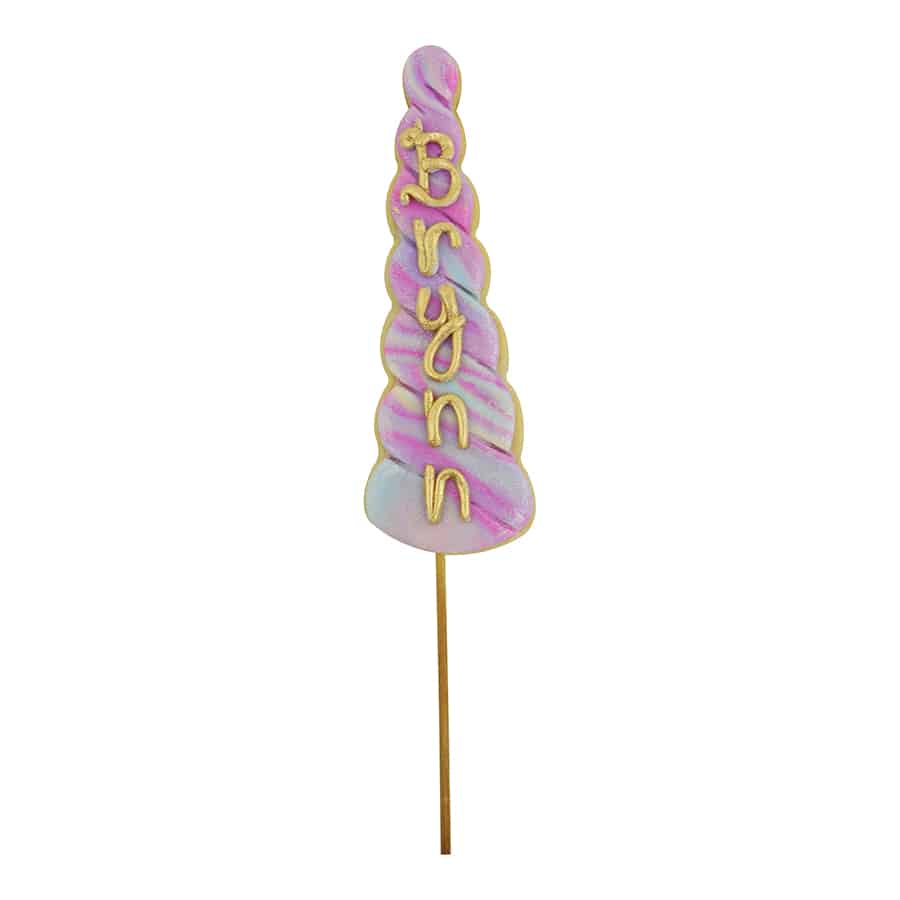 Decorated unicorn horn-shaped cookie with pastel swirls and gold lettering on a wooden stick.