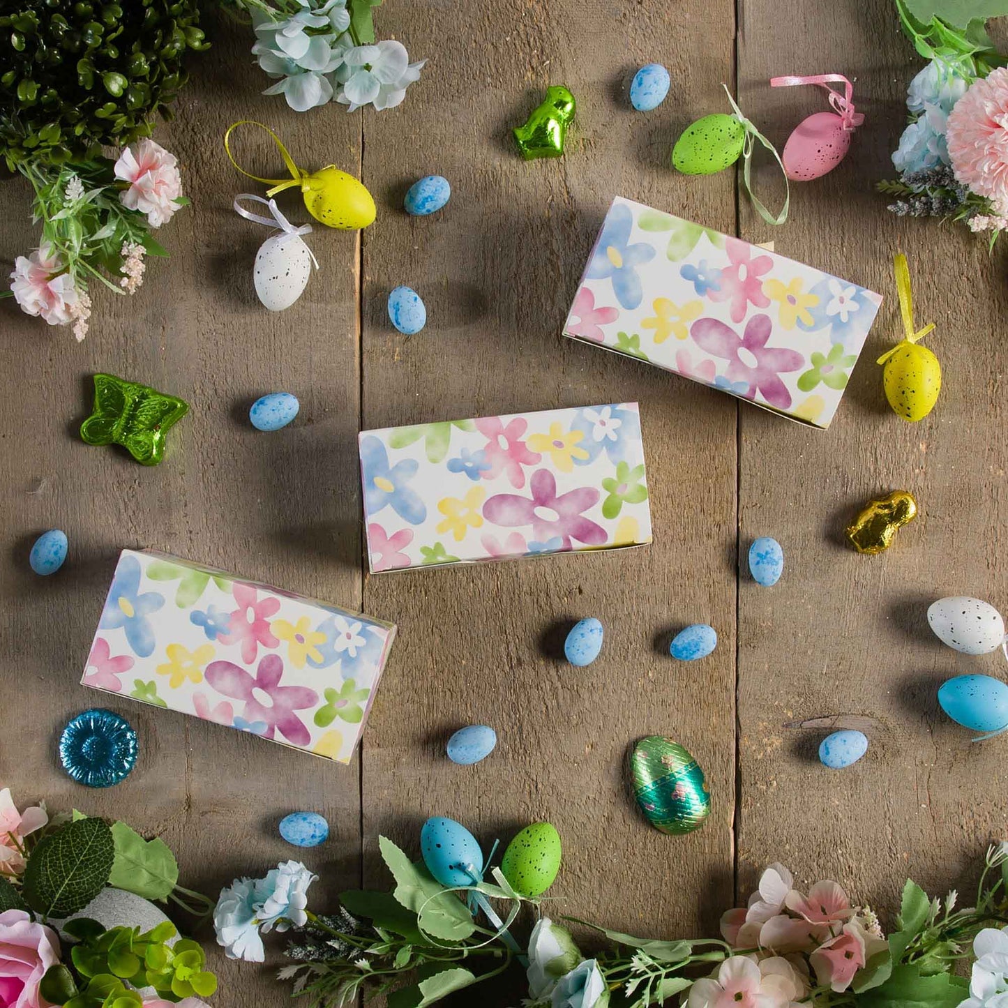 Watercolor daisy candy boxes arranged on a rustic wood surface with spring flowers and decorative Easter eggs.