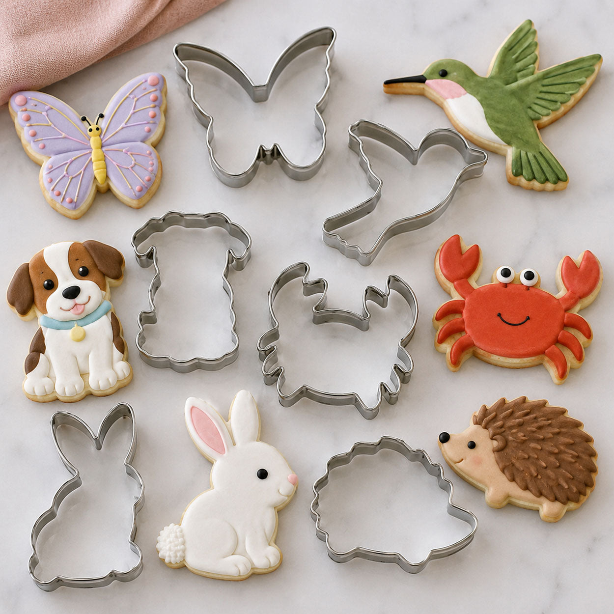 Animal-themed cookie cutters and decorated cookies shaped like a butterfly, hummingbird, puppy, crab, rabbit, and hedgehog on a marble background.