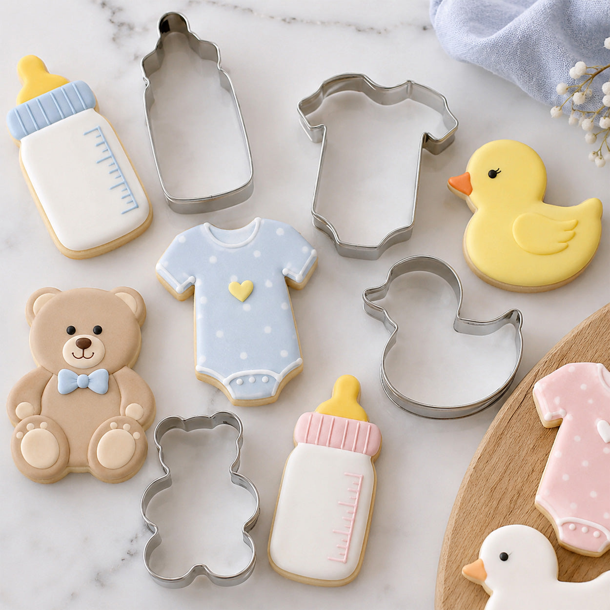 Baby-themed cookie cutters and decorated cookies shaped like baby bottles, onesies, teddy bears, and ducks arranged on a marble surface.