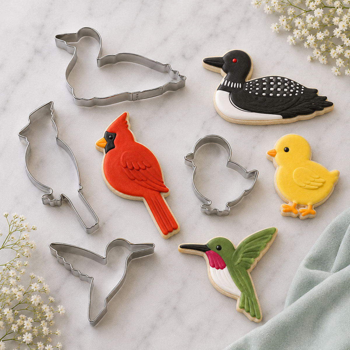 Bird-themed cookie cutters and decorated cookies shaped like a cardinal, hummingbird, duckling, and loon arranged on a marble surface with floral accents.