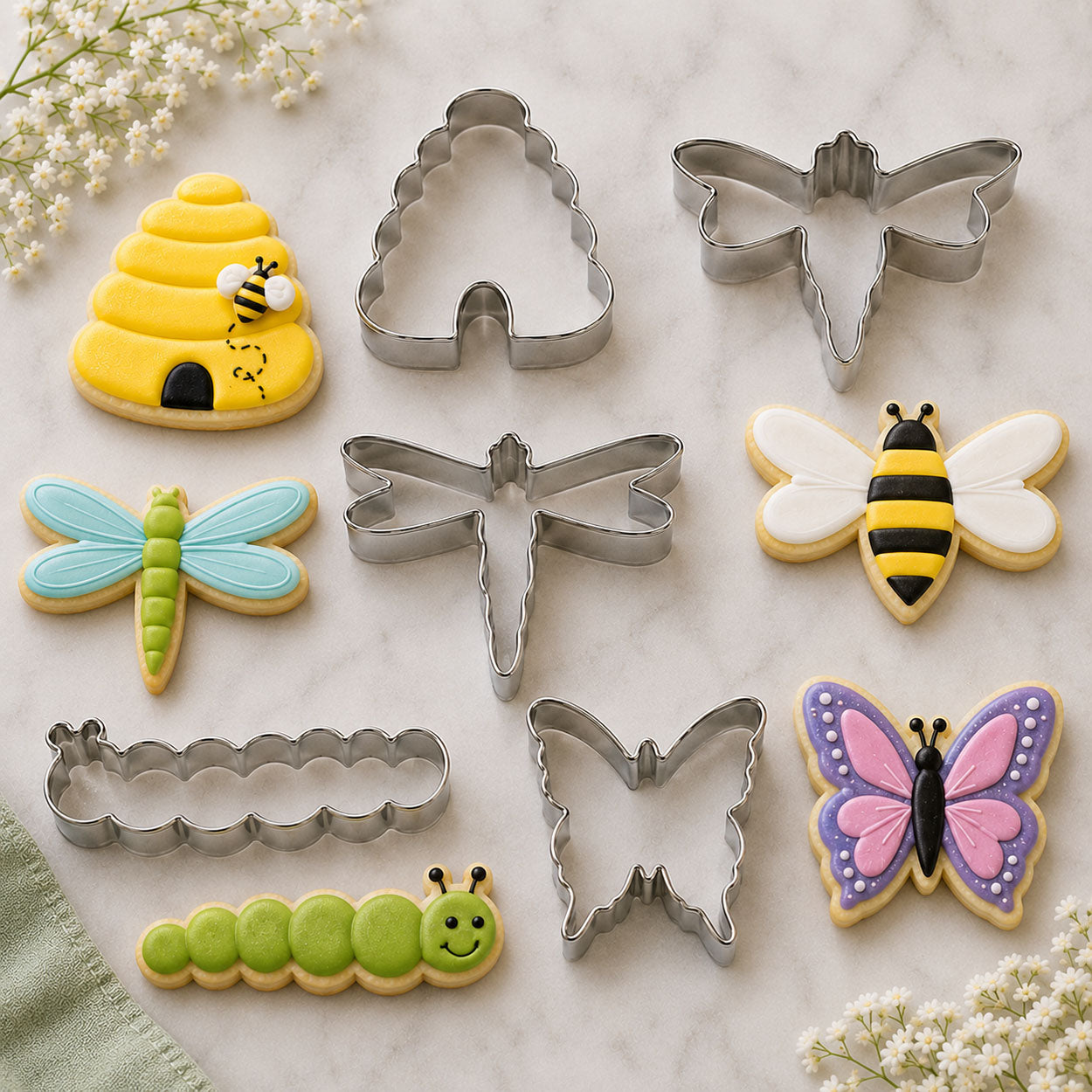 Insect-themed cookie cutters and decorated cookies shaped like a butterfly, bee, dragonfly, caterpillar, and beehive displayed on a marble surface with floral accents.