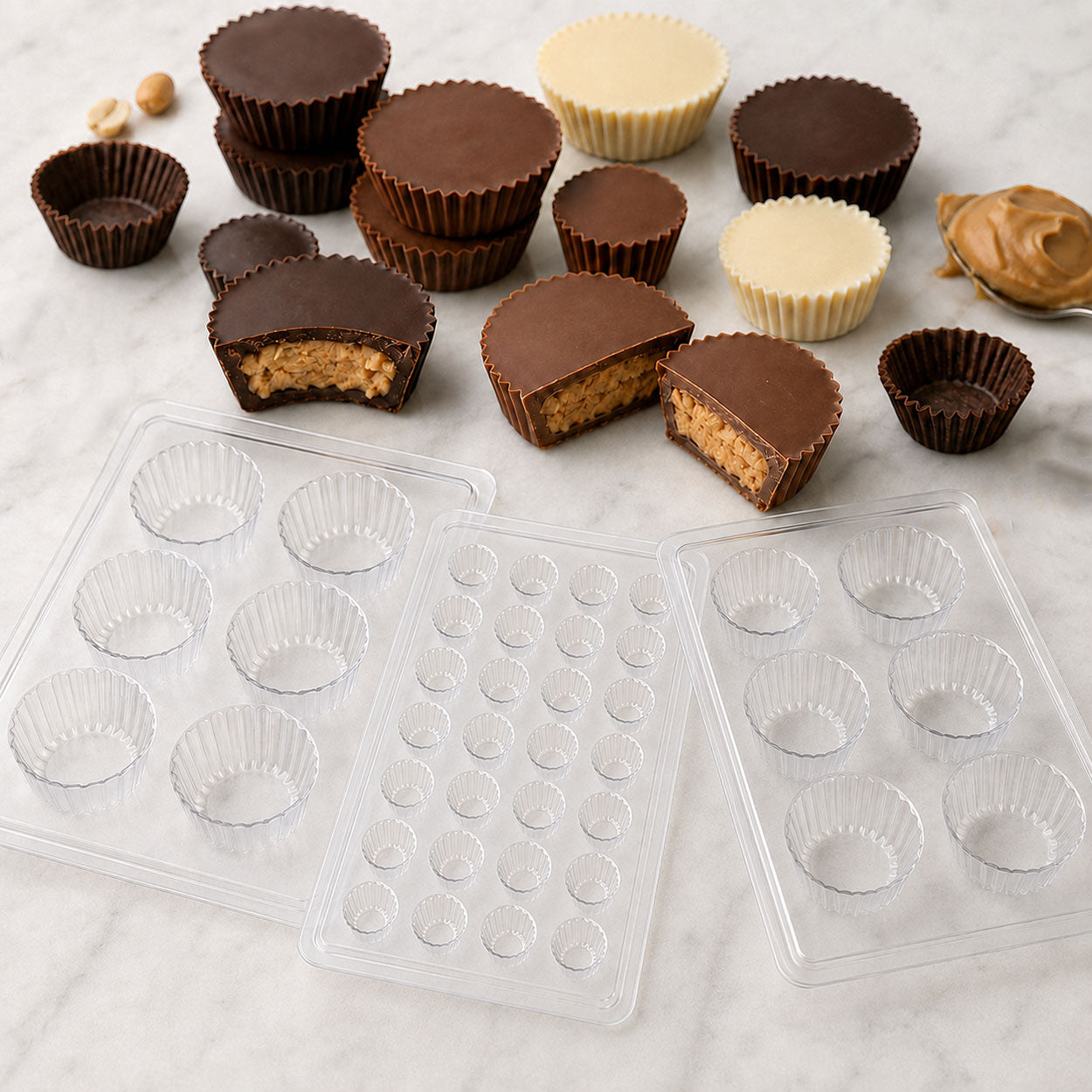 Chocolate peanut butter cups and candy cup molds in assorted sizes with finished milk, dark and white chocolate cups on a marble surface.