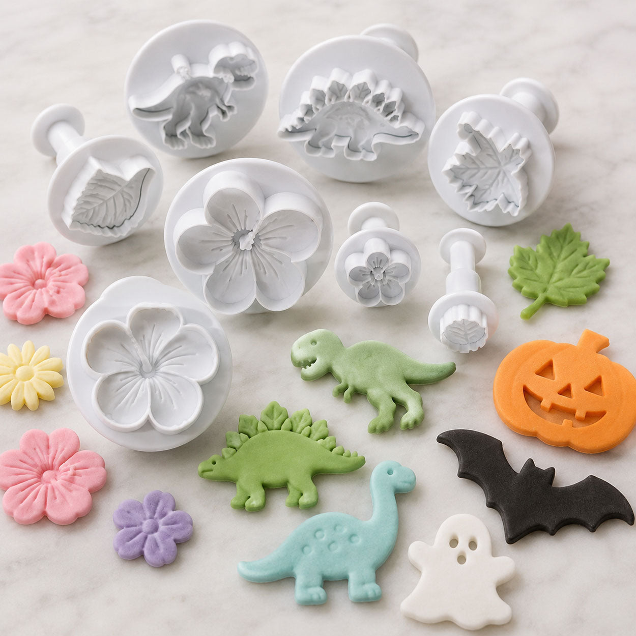 Plunger cookie cutters shaped like dinosaurs, flowers, leaves, and Halloween designs displayed with colorful stamped fondant decorations.