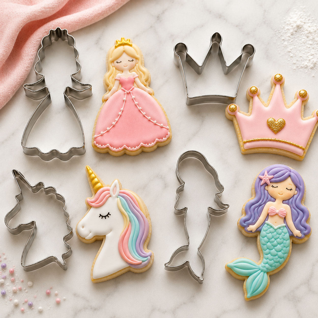 Fantasy cookie cutters and decorated cookies shaped like a princess, crown, unicorn, and mermaid arranged on a marble baking surface.