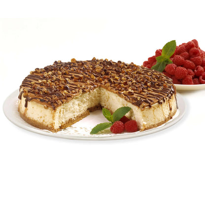 Round cheesecake topped with caramel, chocolate drizzle, and chopped nuts with a slice removed, garnished with raspberries and fresh mint.