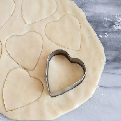 Heart cookie cutter pressing into rolled-out dough with several heart shapes cut out.