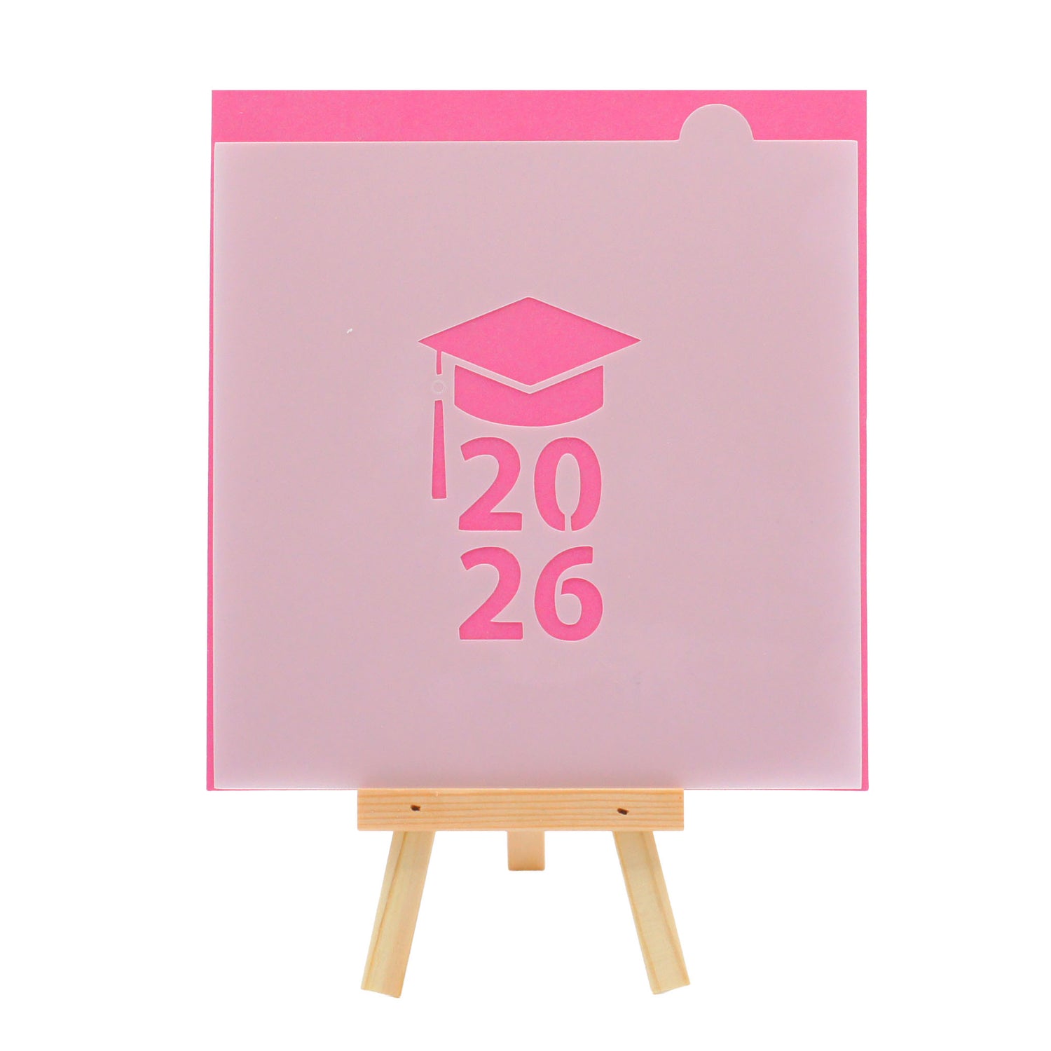 Translucent cookie stencil showing a graduation cap above stacked “2026,” photographed on a small wooden easel against a bright pink background.