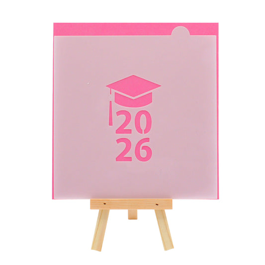 Graduation cap cookie stencil with the year 2026, shown on a bright pink background and displayed on a small wooden easel.