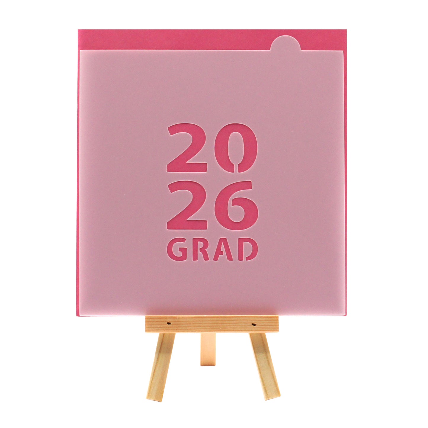 2026 GRAD block lettering cookie stencil on a pink background, displayed on a small wooden easel.