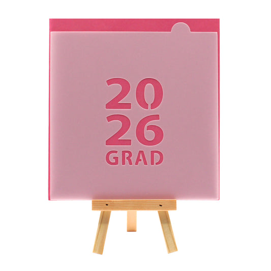 2026 GRAD block lettering cookie stencil on a pink background, displayed on a small wooden easel.