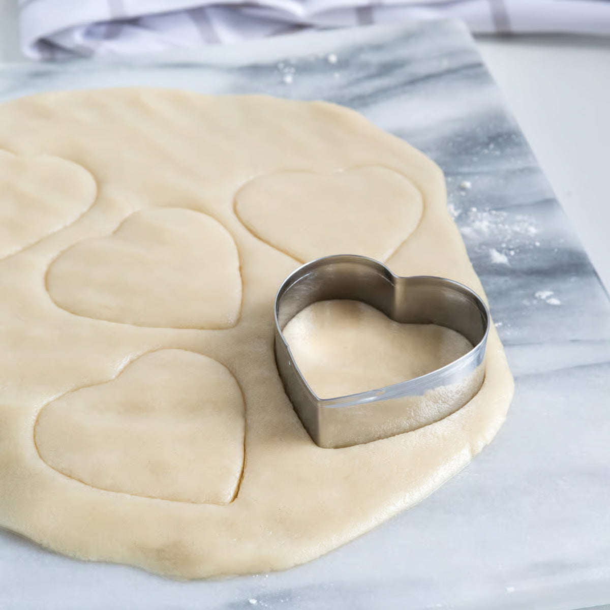 Heart-shaped cookie cutter placed on rolled-out dough with heart shapes already cut out.