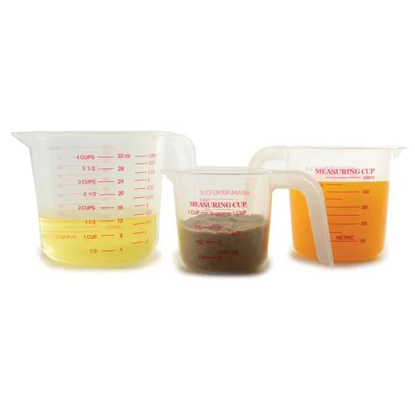 Three transparent plastic measuring cups filled with different colored liquids, each marked with red measurements in cups and milliliters. The largest cup contains yellow liquid, the medium cup contains brown liquid, and the smallest cup contains orange liquid.