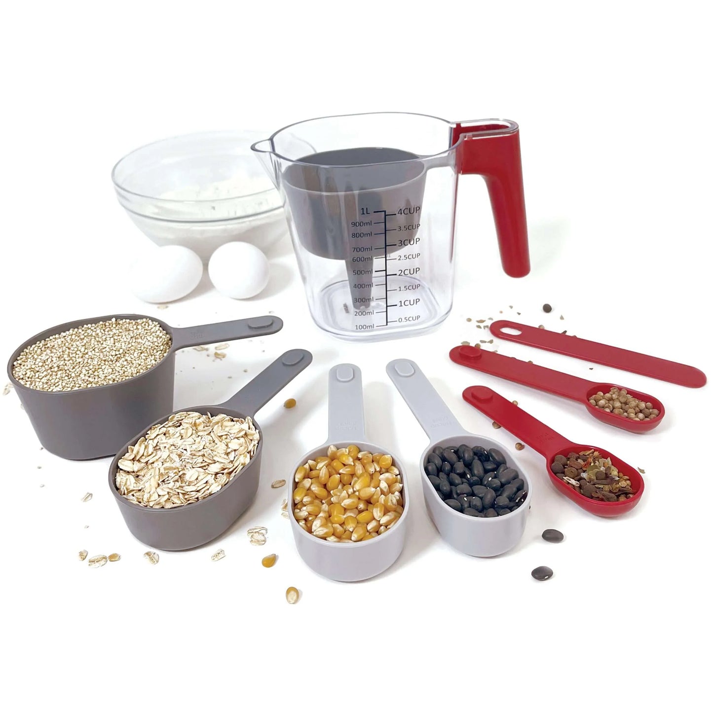 "Set of grey measuring cups and red measuring spoons displayed with various ingredients like oats, quinoa, corn kernels, and black beans. Includes a clear measuring cup with a red handle and a small bowl filled with flour.