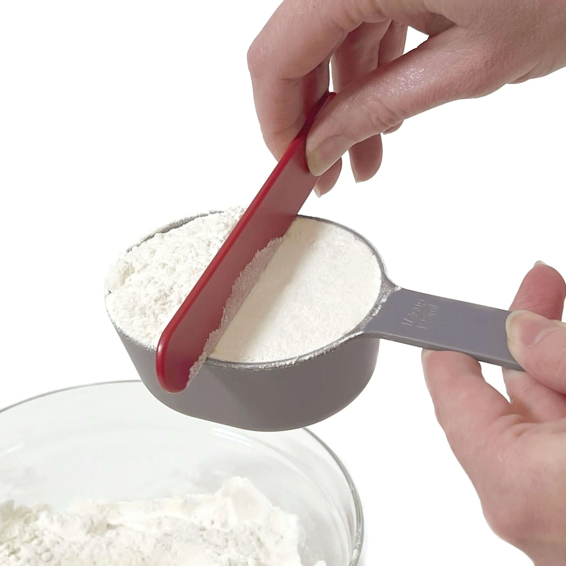 Close-up of a hand using a red plastic leveler to scrape flour from the top of a grey measuring cup filled with flour, ensuring an accurate measurement.