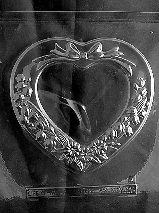 Clear plastic mold showing a heart-shaped chocolate lid with sculpted floral border and bow accent, leaving a smooth center for personalization.