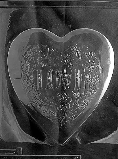 Clear plastic mold featuring a large heart-shaped chocolate lid with an ornate floral design surrounding the embossed word “LOVE” in capital letters.