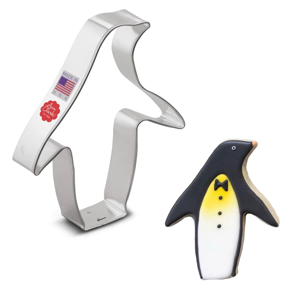 Penguin cookie cutter alongside a decorated penguin-shaped cookie with black, white, and yellow icing, featuring a bow tie and buttons.