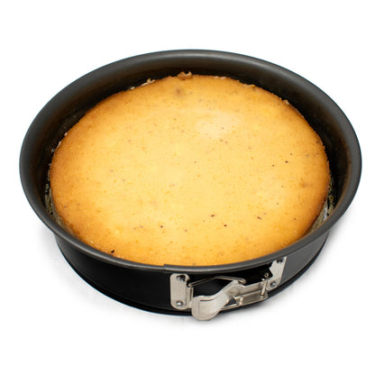 Round golden cheesecake cooling inside a black nonstick springform pan with a visible silver latch on a white background.