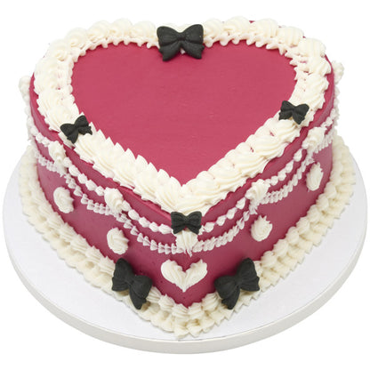 Heart-shaped cake with white frosting, red fondant, and black bow decorations on a white background