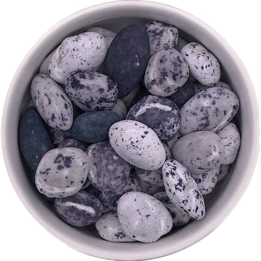 Assorted candy-coated chocolate beach pebbles in gray, blue, and speckled tones, displayed in a white bowl for coastal-themed desserts.