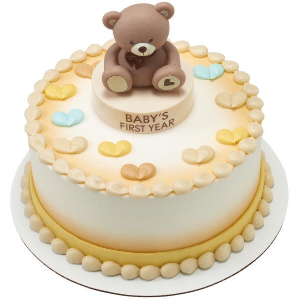 Round celebration cake with soft gradient icing, piped borders, and pastel heart decorations, topped with a brown teddy bear sitting on a “Baby’s First Year” base from the Beary First Year Cake Kit.