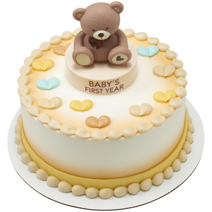 Round celebration cake with soft gradient icing, piped borders, and pastel heart decorations, topped with a brown teddy bear sitting on a “Baby’s First Year” base from the Beary First Year Cake Kit.