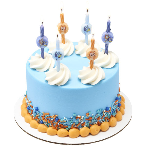 Light blue frosted round cake with white whipped swirls on top, decorated with multiple Bluey character candles and a sprinkle border in blue and orange.