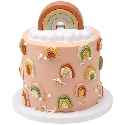 A tall, peach-colored cake decorated with rainbow piping and sprinkles, topped with the stackable rainbow and cloud base.