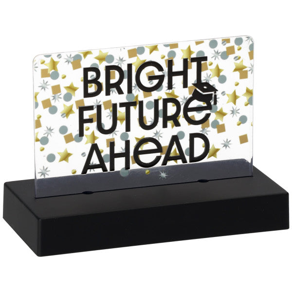 Close-up of a “BRIGHT FUTURE AHEAD” graduation cake topper with black lettering and a small graduation cap icon on a clear plaque with gold and silver confetti shapes, mounted in a black base.