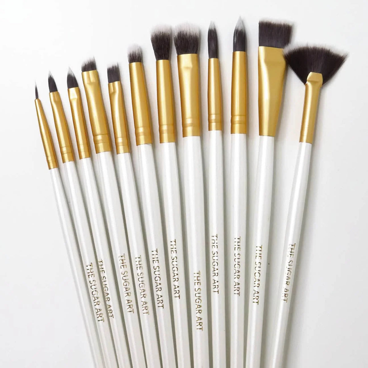 Twelve-piece cake decorating brush set with white handles, gold ferrules, and dark bristles in fine tip, pointed, flat, fluffy, and fan shapes.