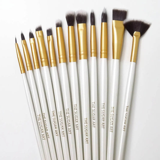 Twelve-piece cake decorating brush set with white handles, gold ferrules, and dark bristles in fine tip, pointed, flat, fluffy, and fan shapes.