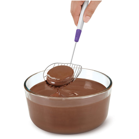 Hand using Wilton Candy Melt Dipping Scoop to lift a chocolate-covered treat from a bowl of melted candy coating.