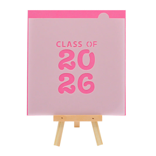 Translucent graduation cookie stencil with the words “CLASS OF 2026” in bold block text, displayed on a small wooden easel against a bright pink background.