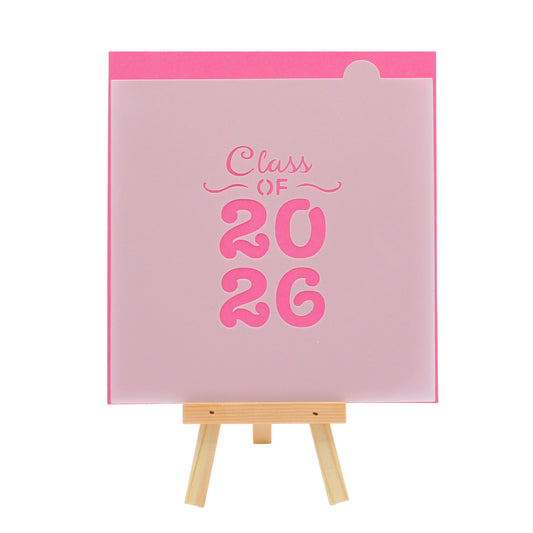 Translucent graduation cookie stencil featuring the words “Class of 2026” in script and bold block numbers, displayed on a small wooden easel against a bright pink background.