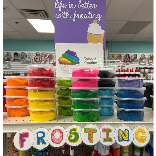 A tantalizing selection of vividly colored buttercream frosting, presented in transparent containers neatly stacked in a vibrant rainbow gradient. Situated in a well-lit baking supplies store, the frosting collection sits beneath an inviting purple sign with a whimsical cupcake graphic proclaiming 'life is better with frosting'. Above the eye-catching display, the word 'FROSTING' is spelled out in colorful, frosted lettering, signaling the rich palette of flavors and creative possibilities