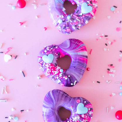 Heart-shaped donuts with vibrant purple and pink marbled glaze decorated with pastel blue heart candies and colorful sprinkles
