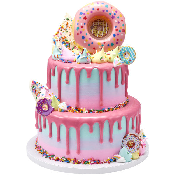 A two-tiered pastel-colored drip cake topped with a large donut and surrounded by mini donuts, sprinkles, and meringue decorations.