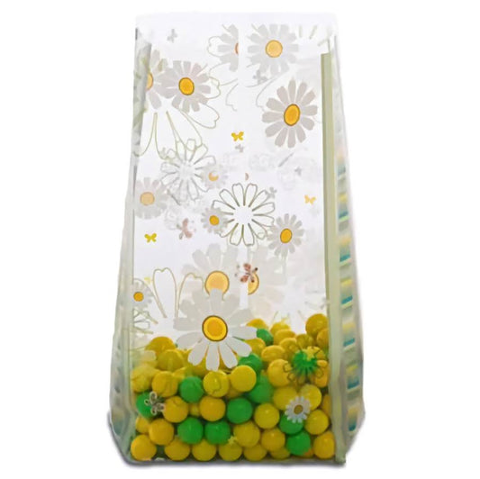 A medium-sized cellophane treat bag filled with bright yellow and green candies at the bottom. The bag is adorned with a delicate pattern of white daisies with yellow centers, and a few small bees scattered throughout. The transparent design with flowers creates a cheerful, spring-themed appearance, perfect for parties or as a sweet gift.