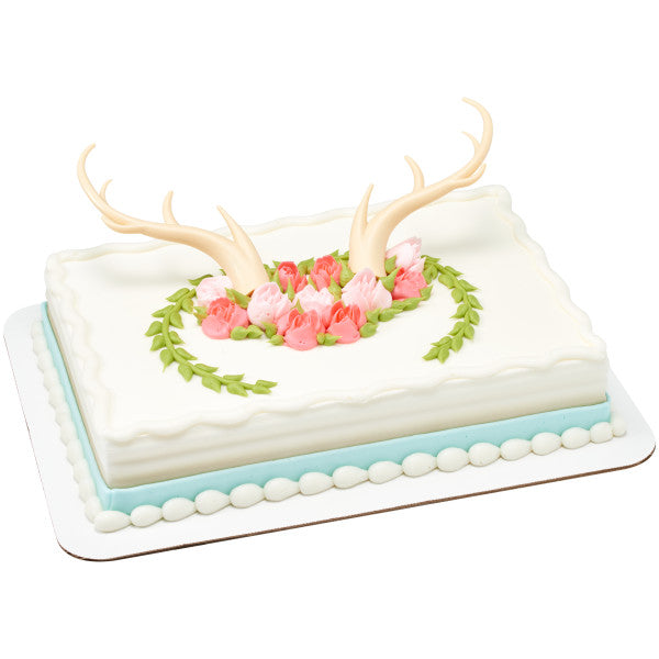 Rustic antler cake topper set on a white rectangular cake decorated with pink flowers and green leaves.