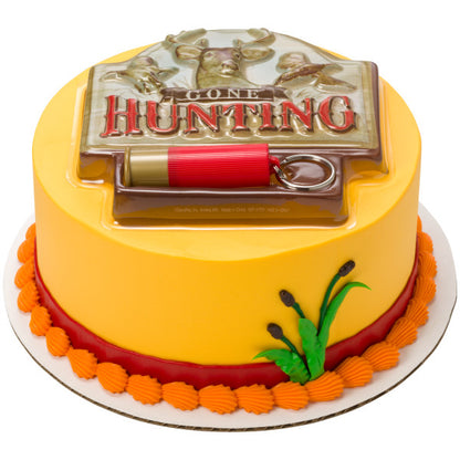 Round yellow cake with autumn-inspired colors, green icing foliage, and a Gone Hunting cake topper plaque with red cartridge shell keychain.